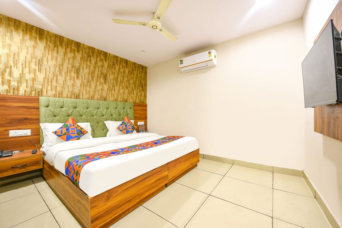 A bedroom with a green tufted headboard, a bed with white bedding and colorful pillows, a wooden nightstand, a bedside table with a phone and remote, a ceiling fan, and an air conditioning unit.