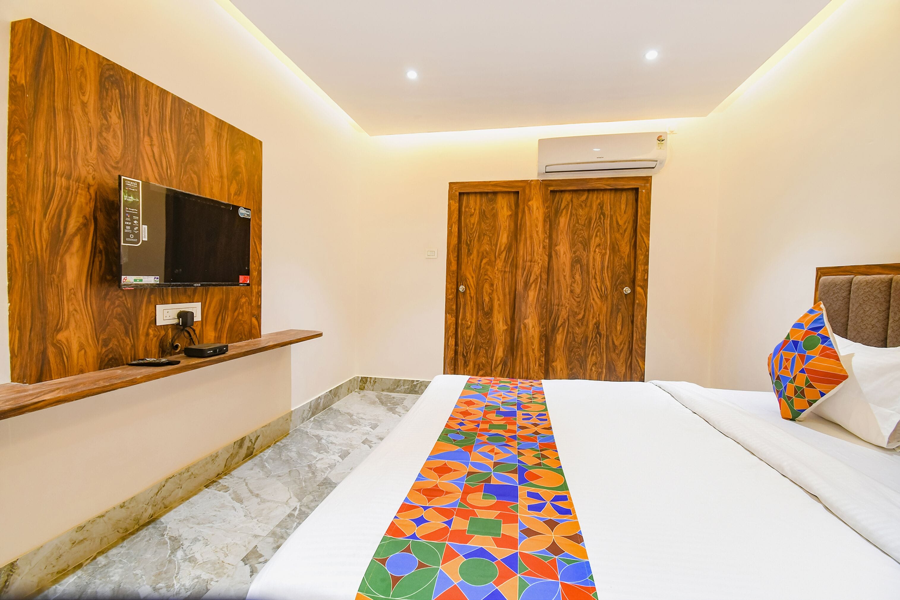 Une chambre d’hôtel avec un mur en bois, un téléviseur fixé au mur, un lit recouvert d’une couverture aux motifs géométriques colorés et une porte en bois.