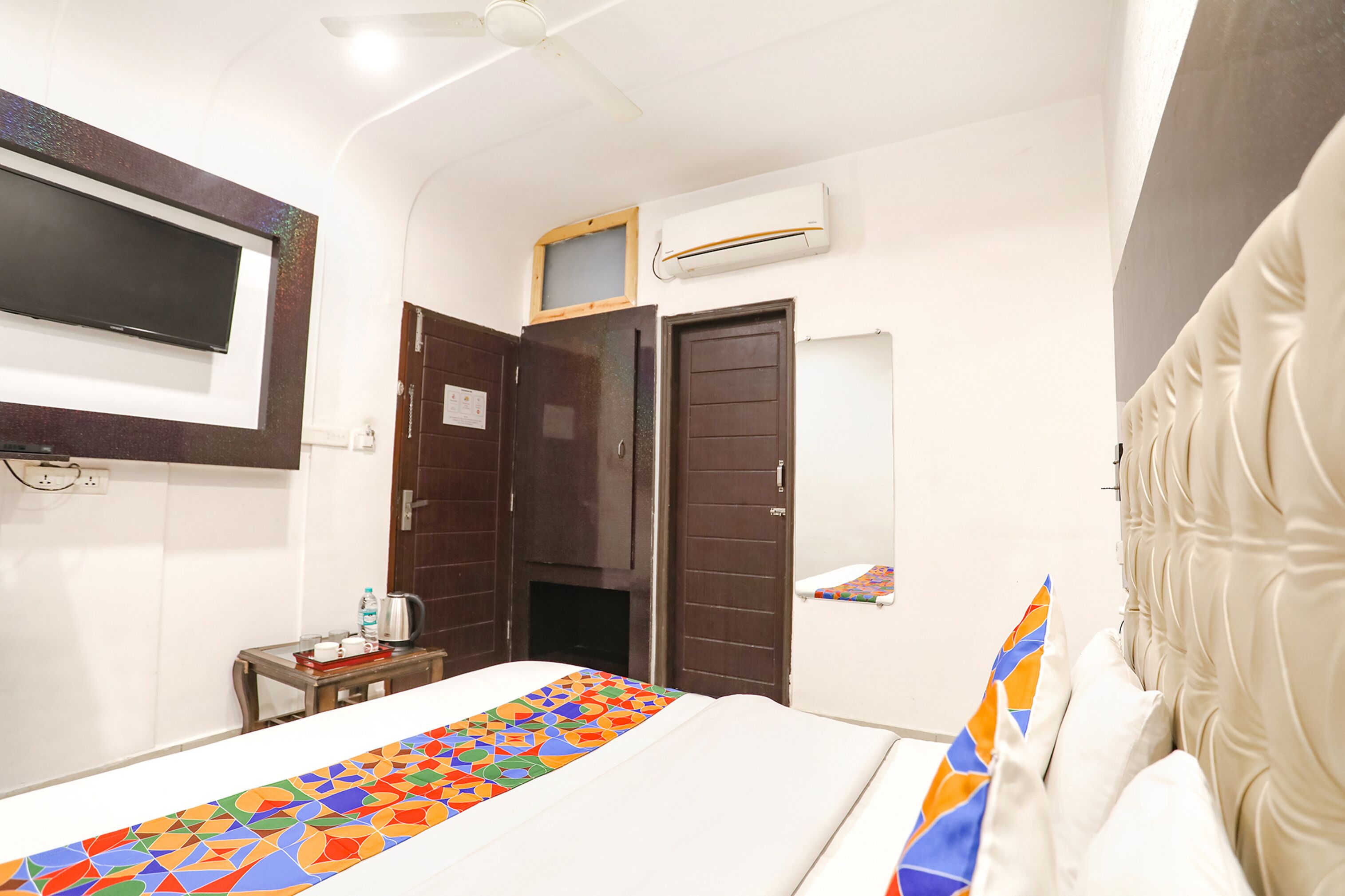 A hotel room with a bed, a TV mounted on the wall, an air conditioning unit, and a small table with a kettle and a bottle.