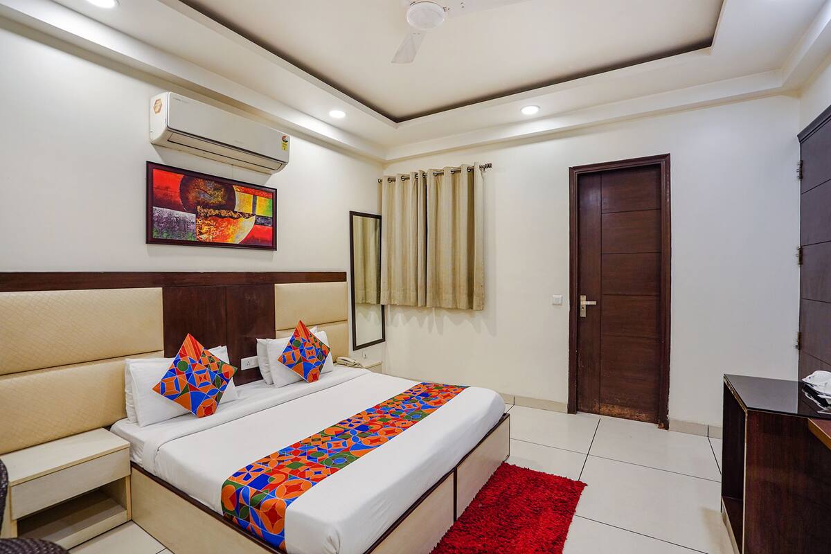 A hotel room with a bed, bedside table, air conditioning unit, artwork, curtains, and a door.