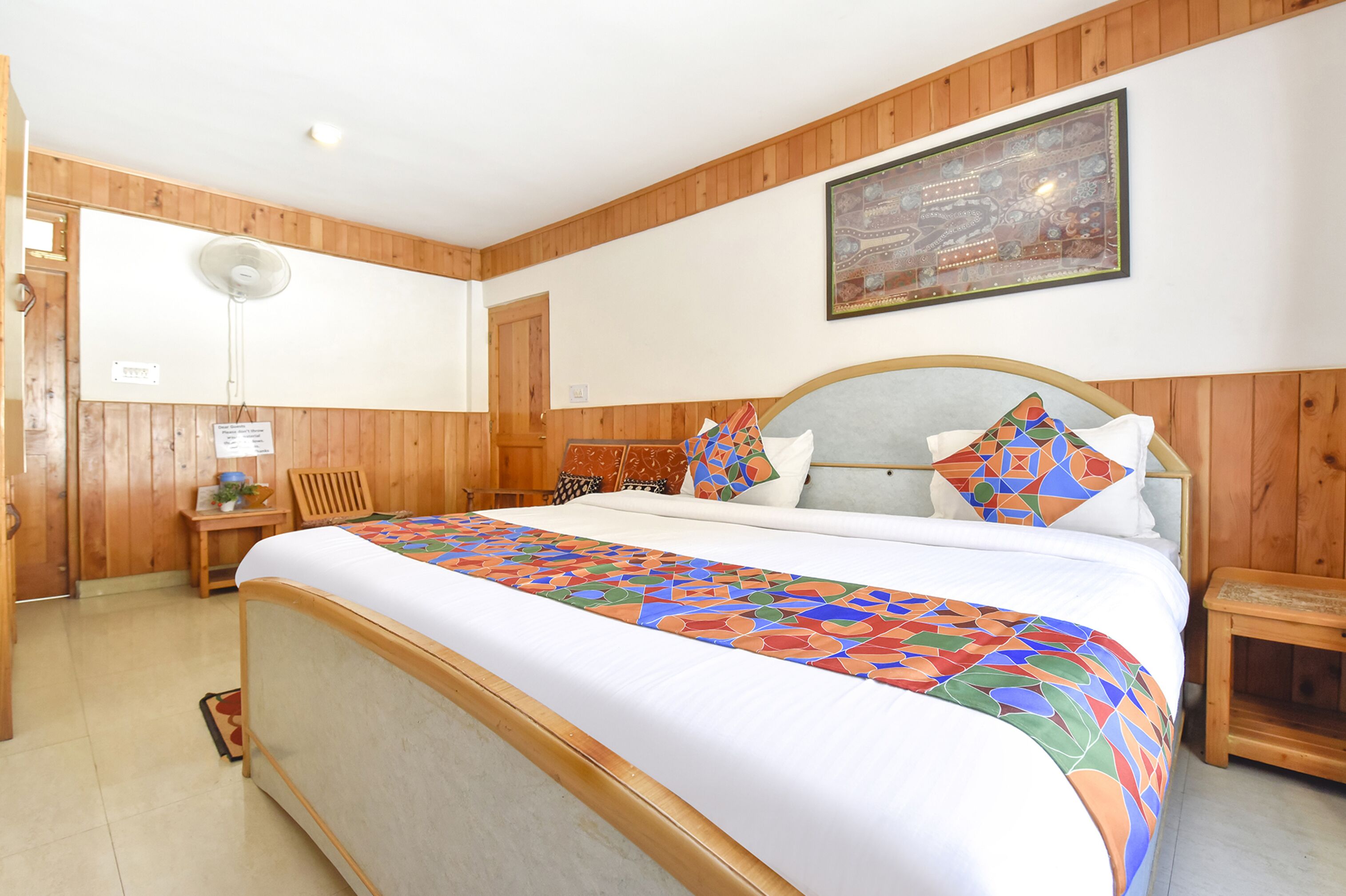 A bedroom with a large bed, wooden panel walls, a ceiling fan, a small table with a plant, and a framed picture on the wall.