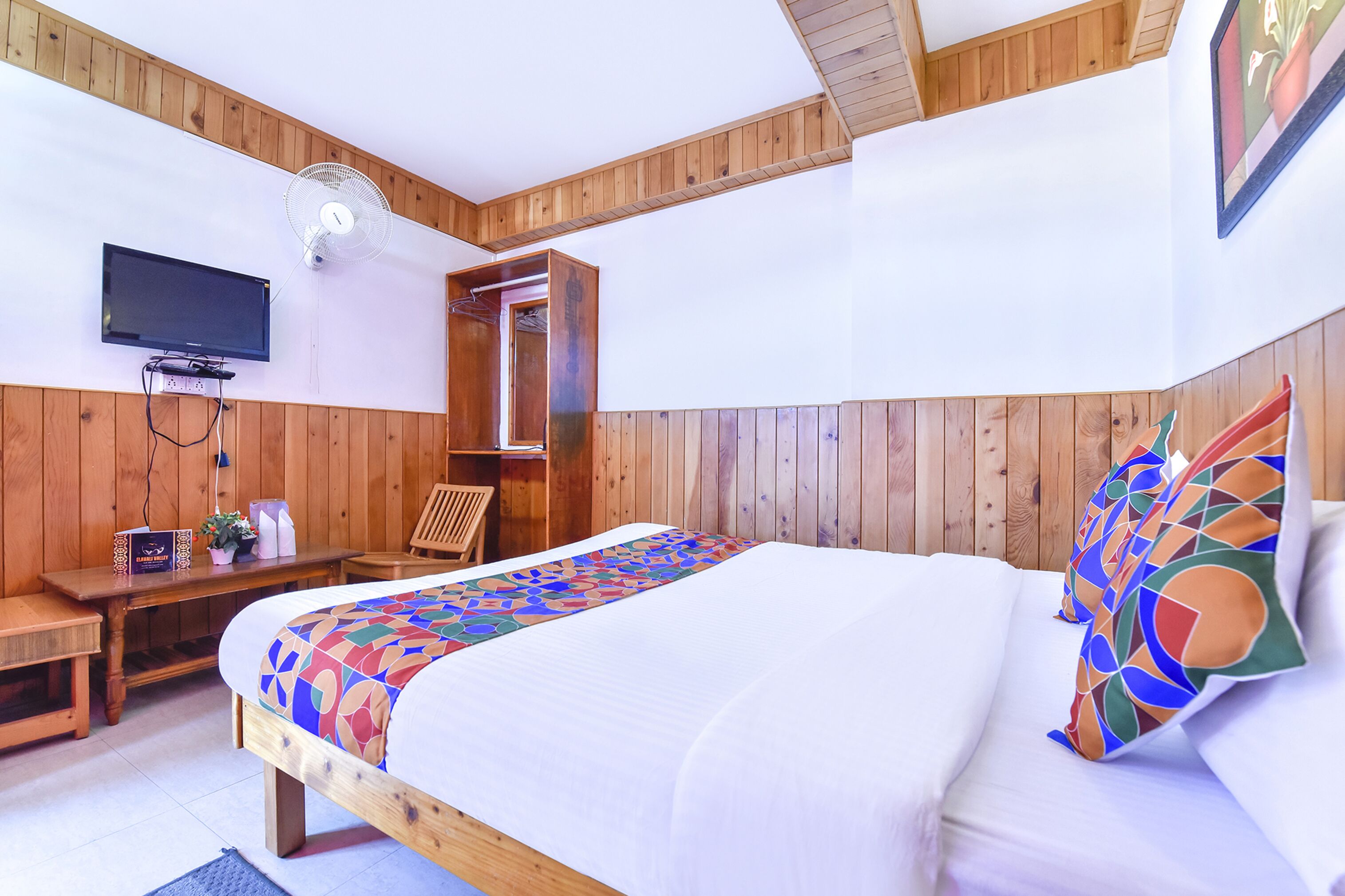 A bedroom with a wooden headboard, a bed with a colorful geometric-patterned bedspread, a bedside table with a lamp, a chair, and a TV mounted on the wall.