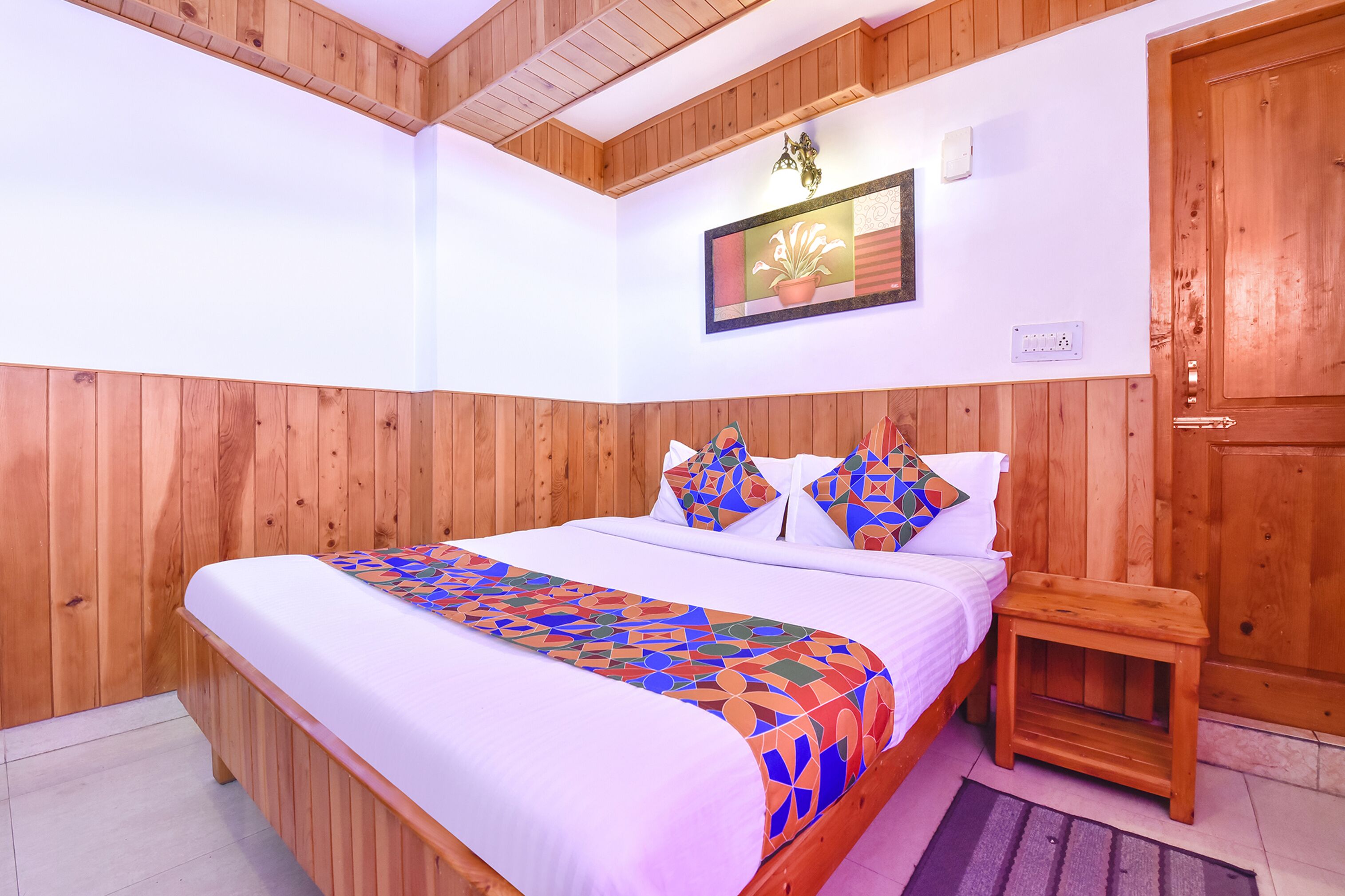 Dormitorio con paneles de madera, una cama, almohadas, un taburete pequeño de madera y una lámpara de pared.