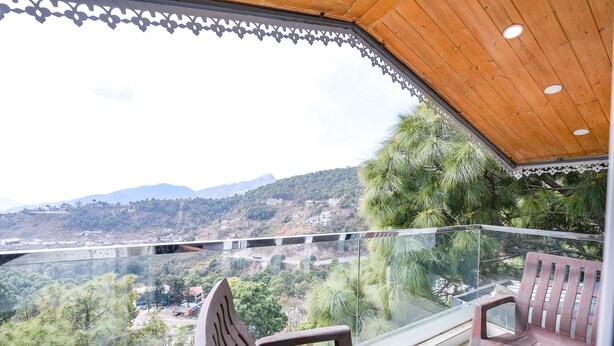 Un balcon doté d’un plafond en bois et d’une balustrade en verre, offrant une vue sur un paysage montagneux parsemé d’arbres et de bâtiments.