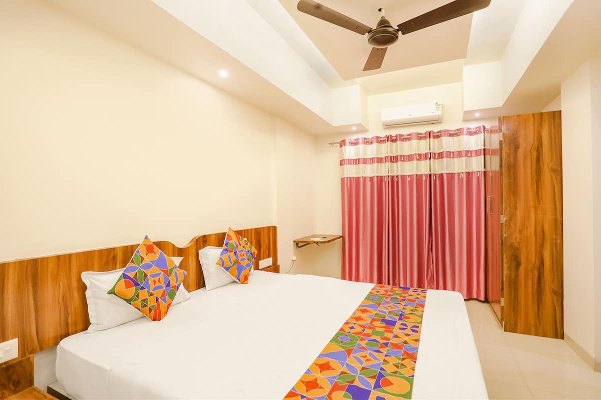 Quarto de hotel com cama, cabeceira de madeira, ventoinha no teto, aparelho de ar condicionado e janela com cortinas.