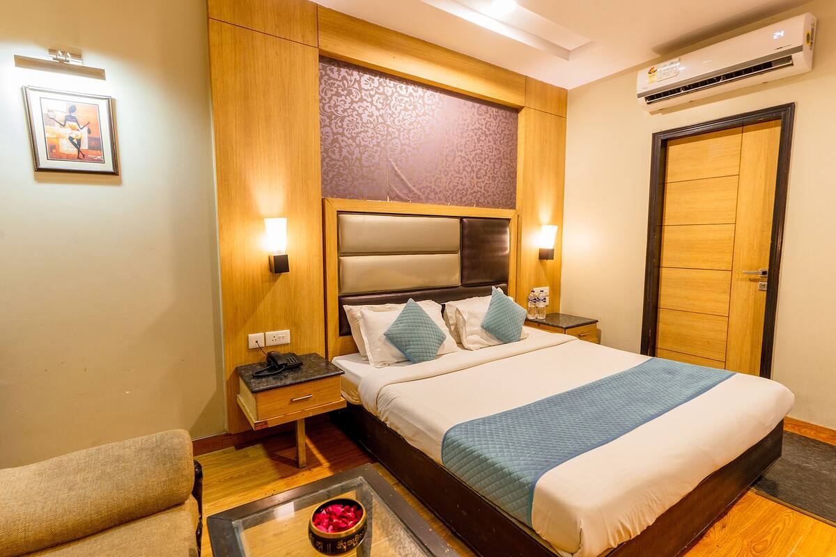 Quarto de hotel com uma cama grande, cabeceira de madeira, duas mesas de cabeceira com luminárias, uma pequena mesa com um objeto decorativo e uma porta com moldura de madeira.