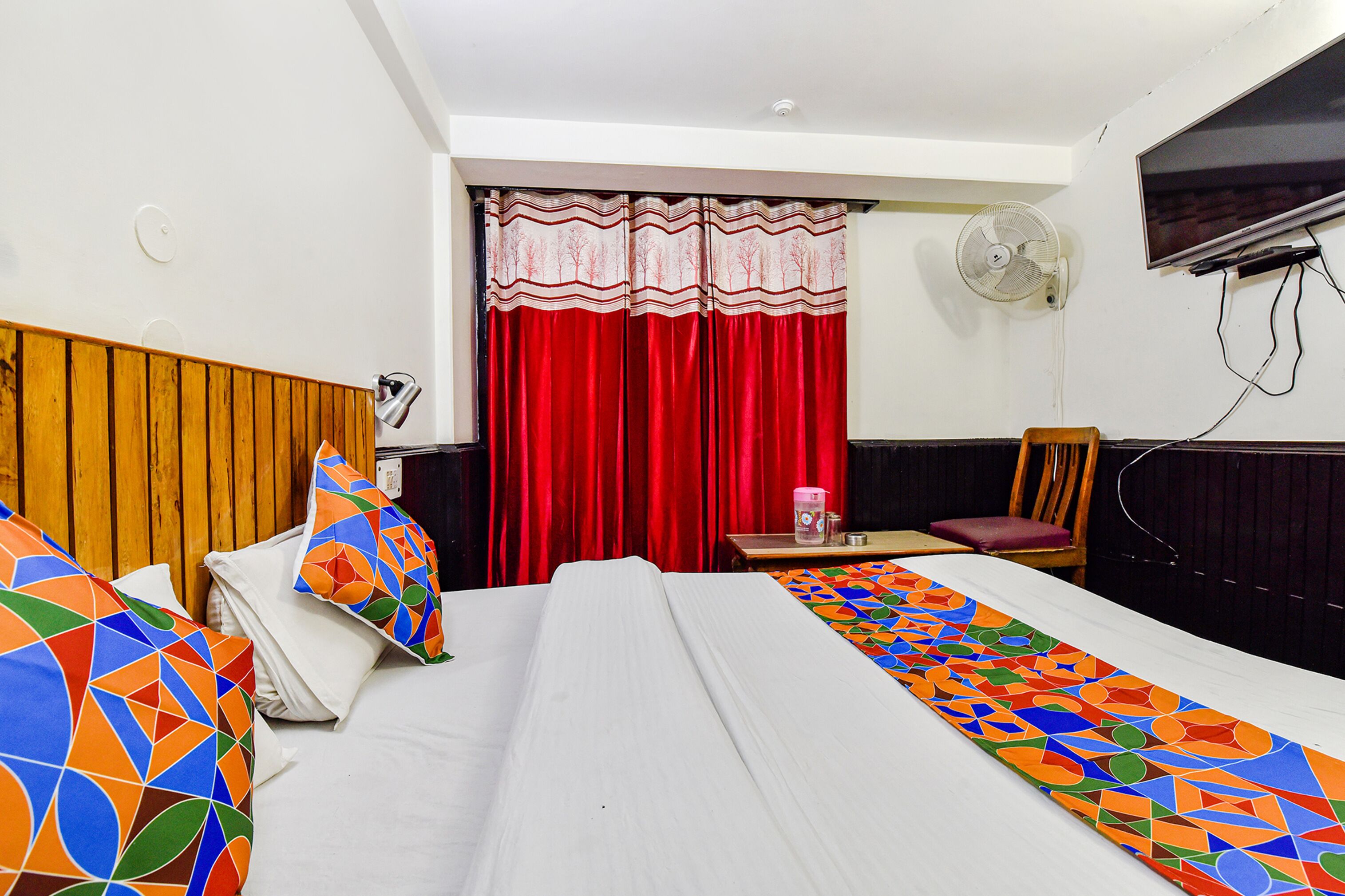 A hotel room with a bed, a wooden headboard, a desk with a chair, a TV, and a window with red curtains.