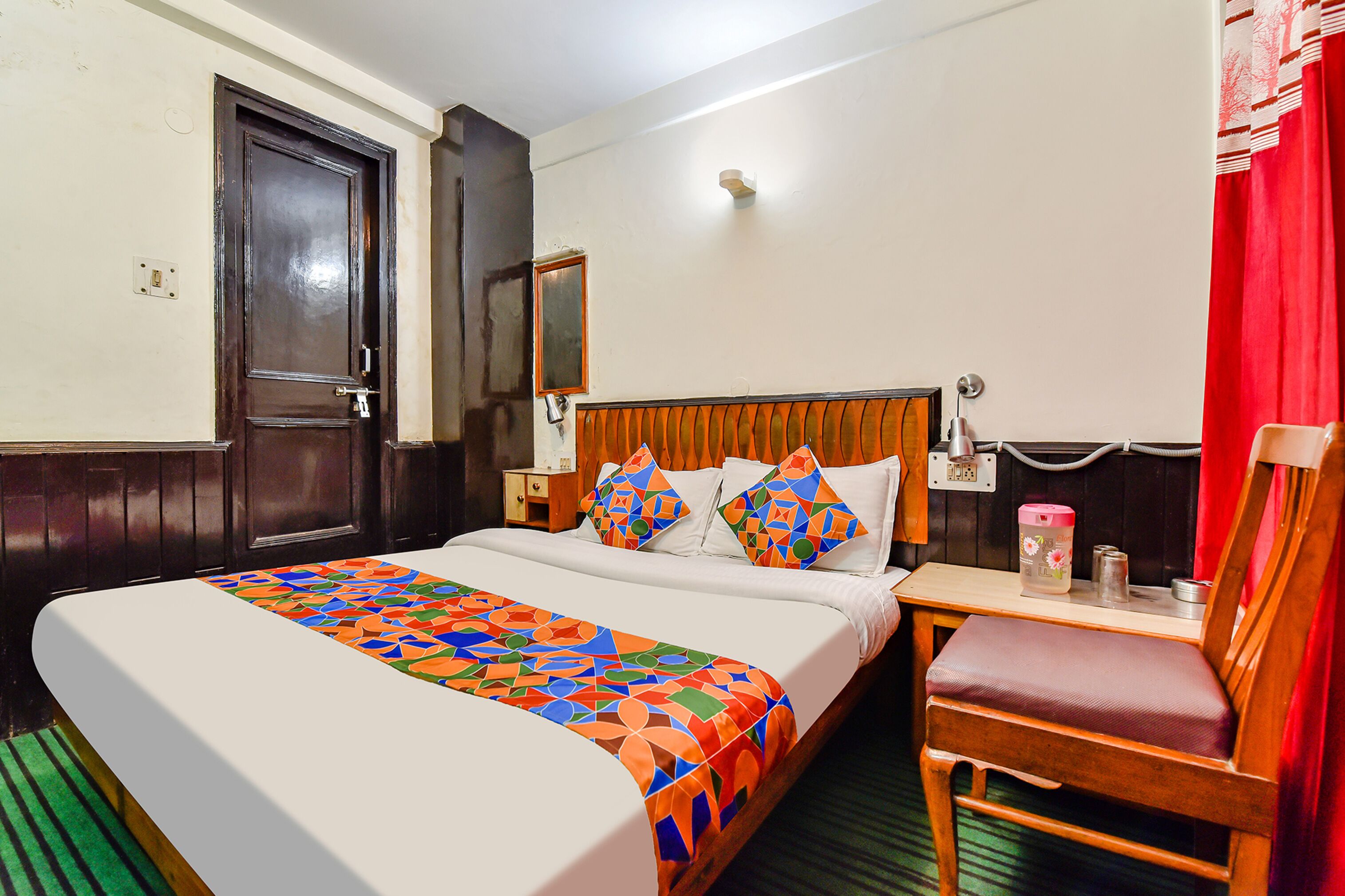 A hotel room with a large bed, a wooden headboard, a chair, a nightstand, and a red curtain.