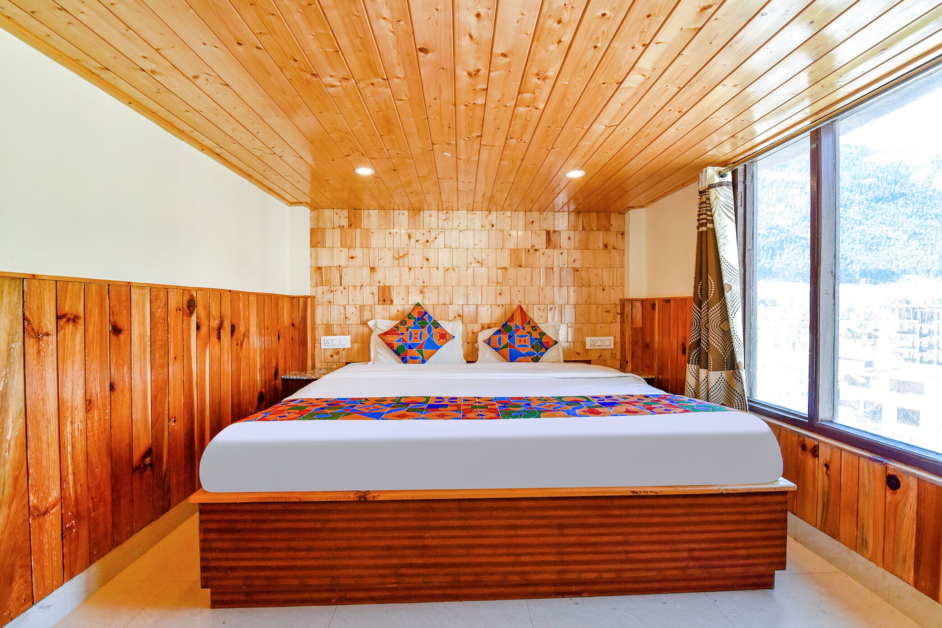 Une chambre à coucher aux murs lambrissés, avec un grand lit, une fenêtre donnant sur des immeubles et une couvre-lit à motifs.