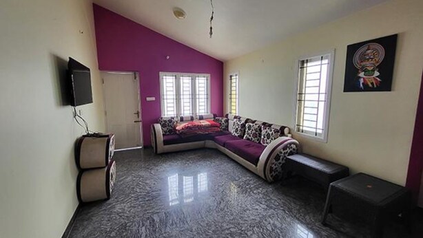 A room with a curved sofa, a television mounted on the wall, and a window with blinds.