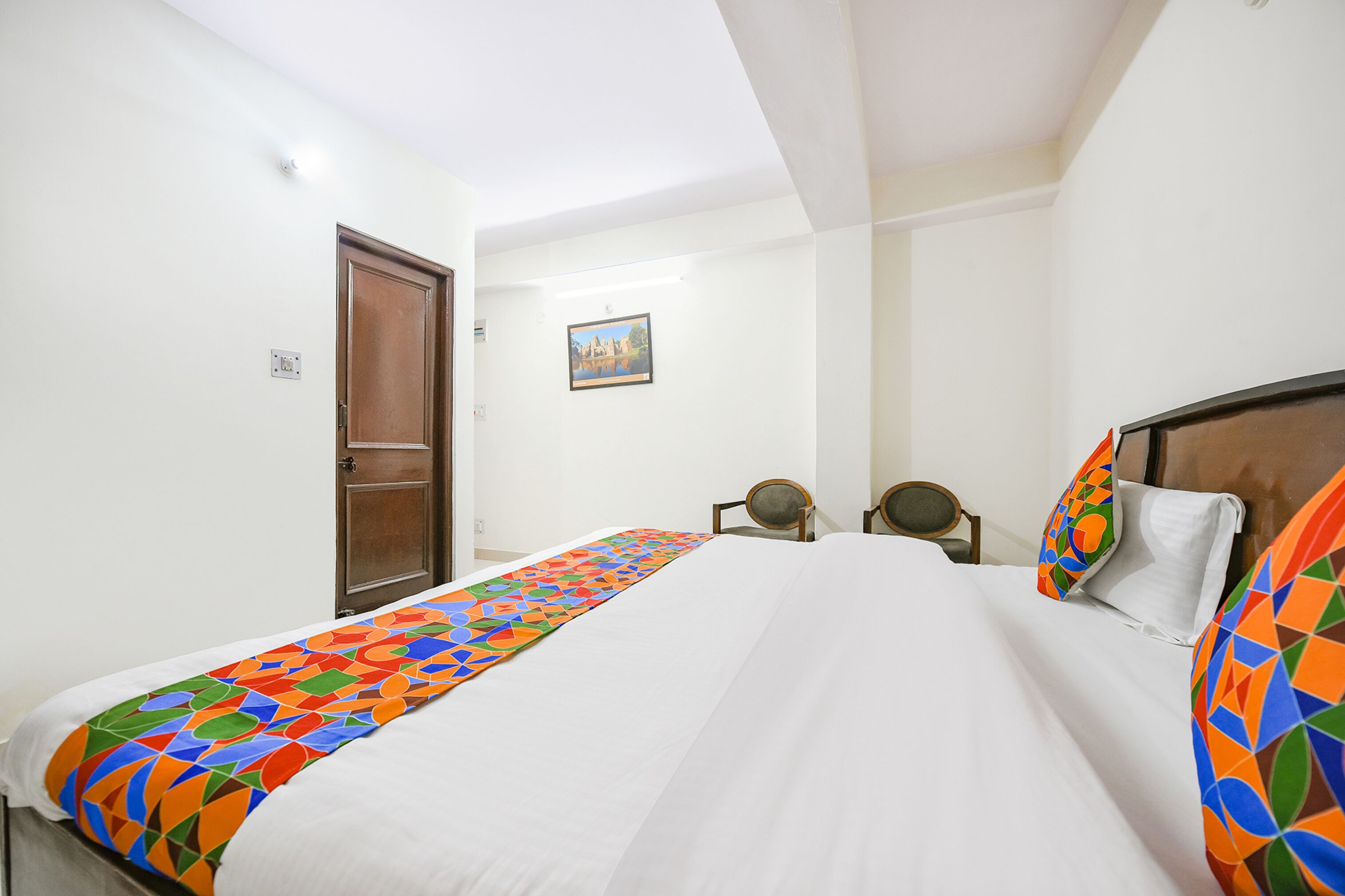 Una habitación de hotel con cama, una puerta de madera, una mesa redonda pequeña y un cuadro colgado en la pared.