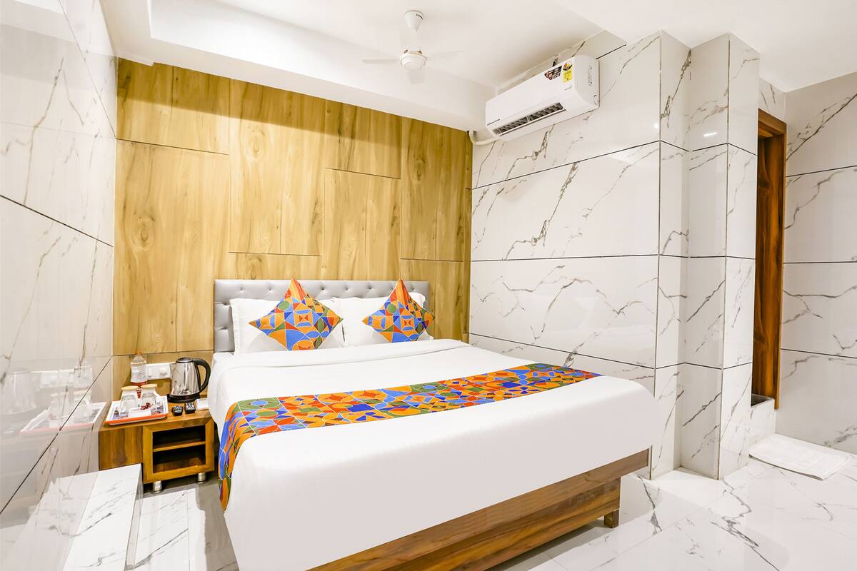 A modern hotel room with a wooden headboard, a large bed with a colorful geometric-patterned bedspread, a bedside table with a coffee maker, and marble-patterned walls.