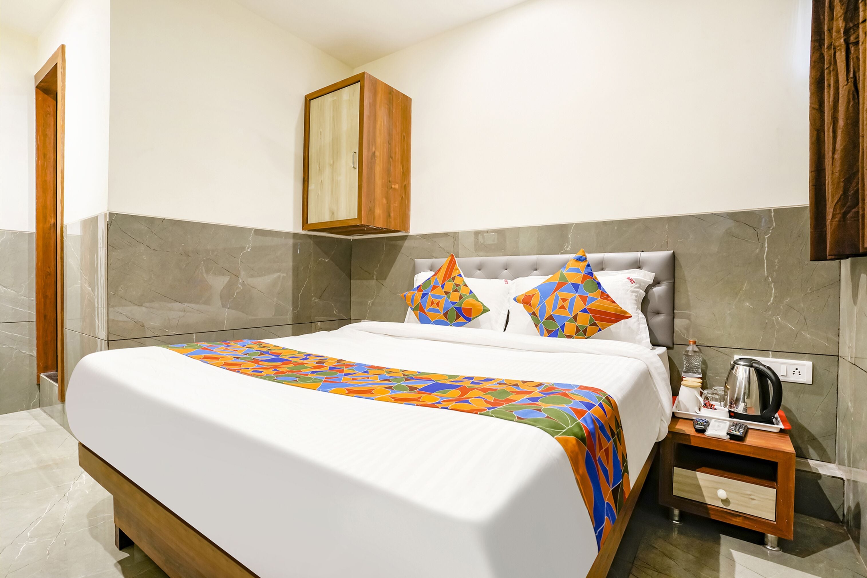 A neatly made bed with a colorful geometric-patterned bedspread, a grey upholstered headboard, and a wooden nightstand with a kettle and a glass of water.