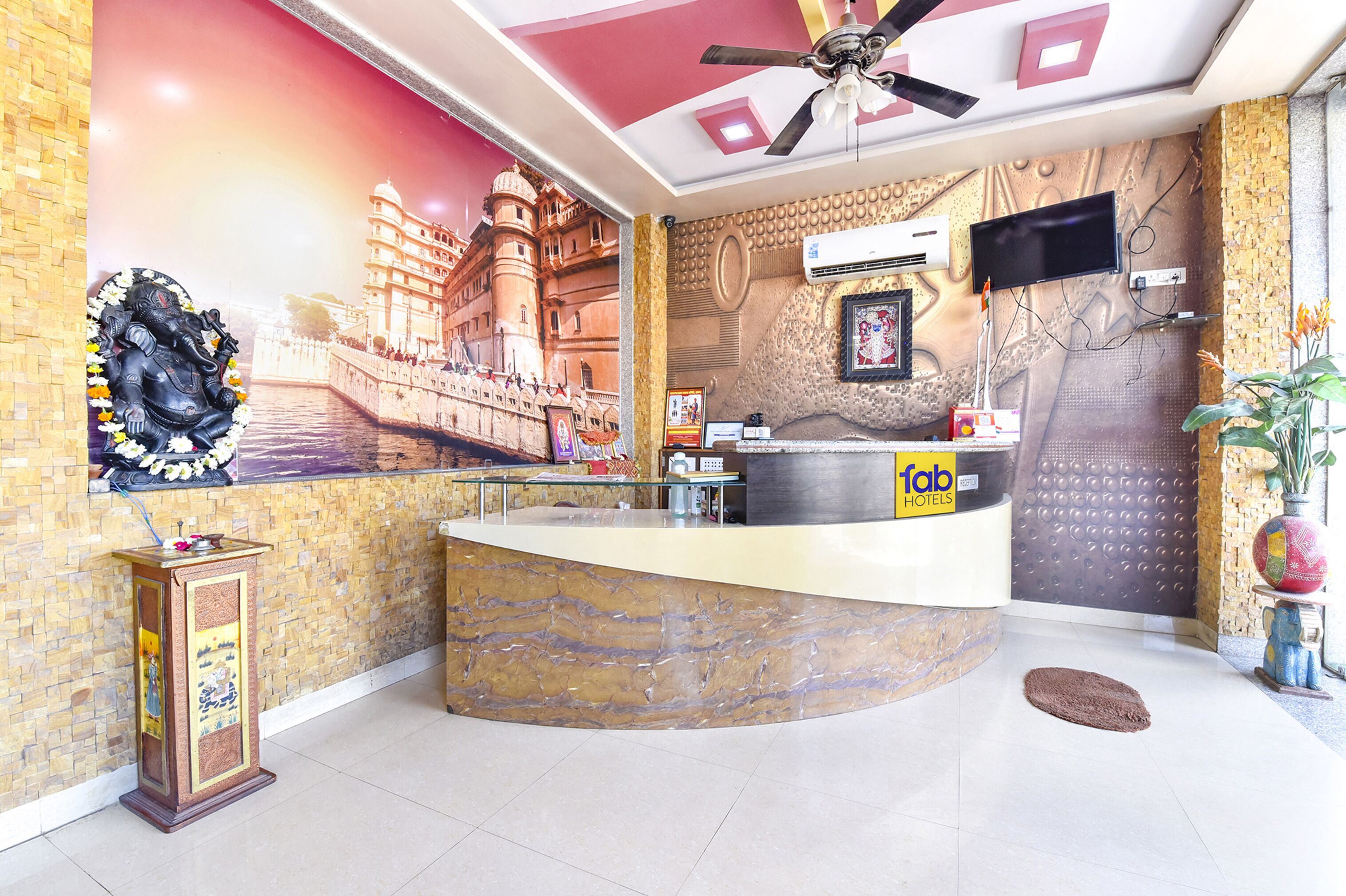 Zona de recepción de hotel con mostrador curvo, ventilador de techo, televisor montado en la pared y un mural que representa un edificio histórico.