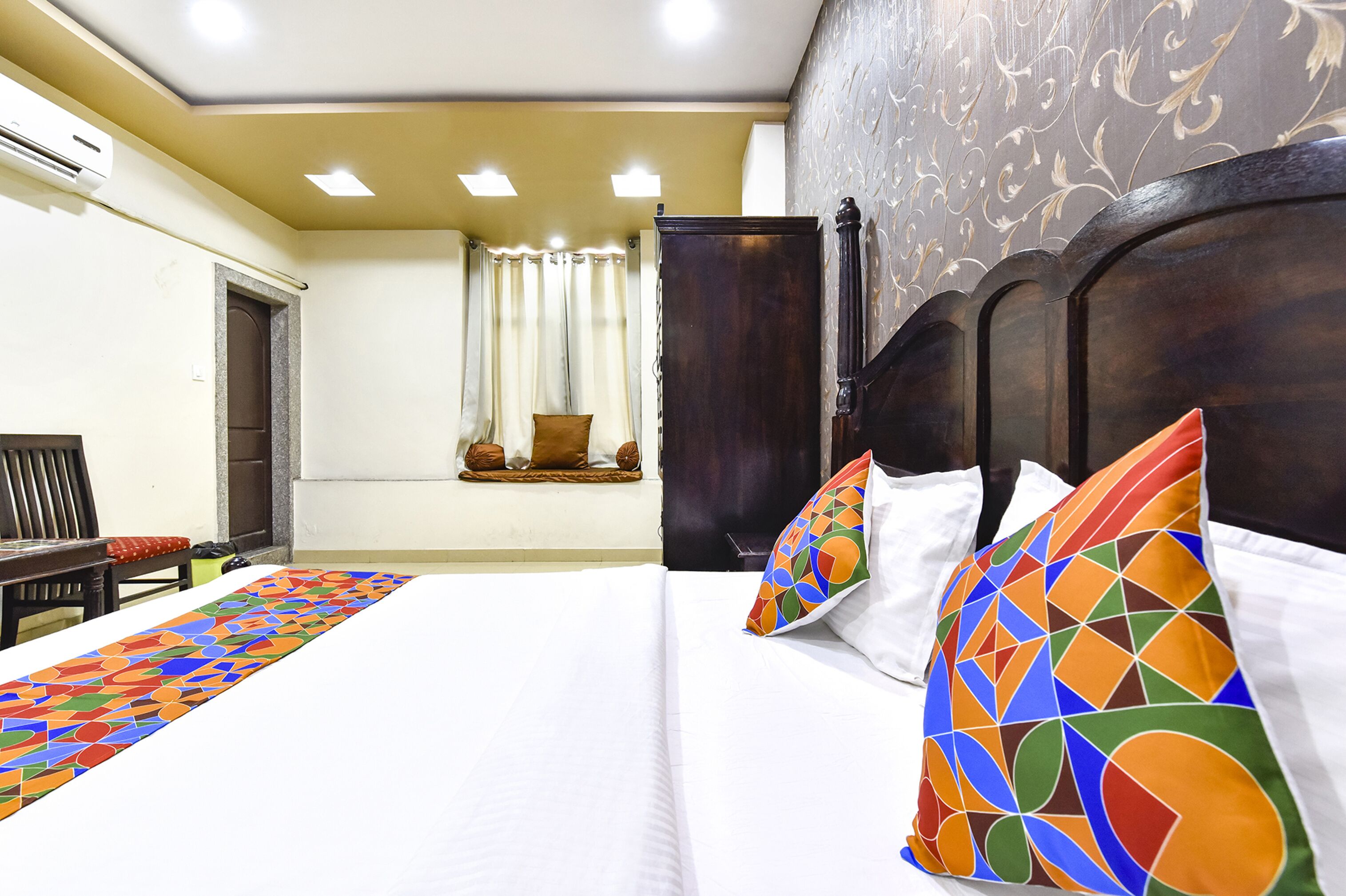 A hotel room with a large bed, a wooden headboard, a window with curtains, and a small table with a chair.