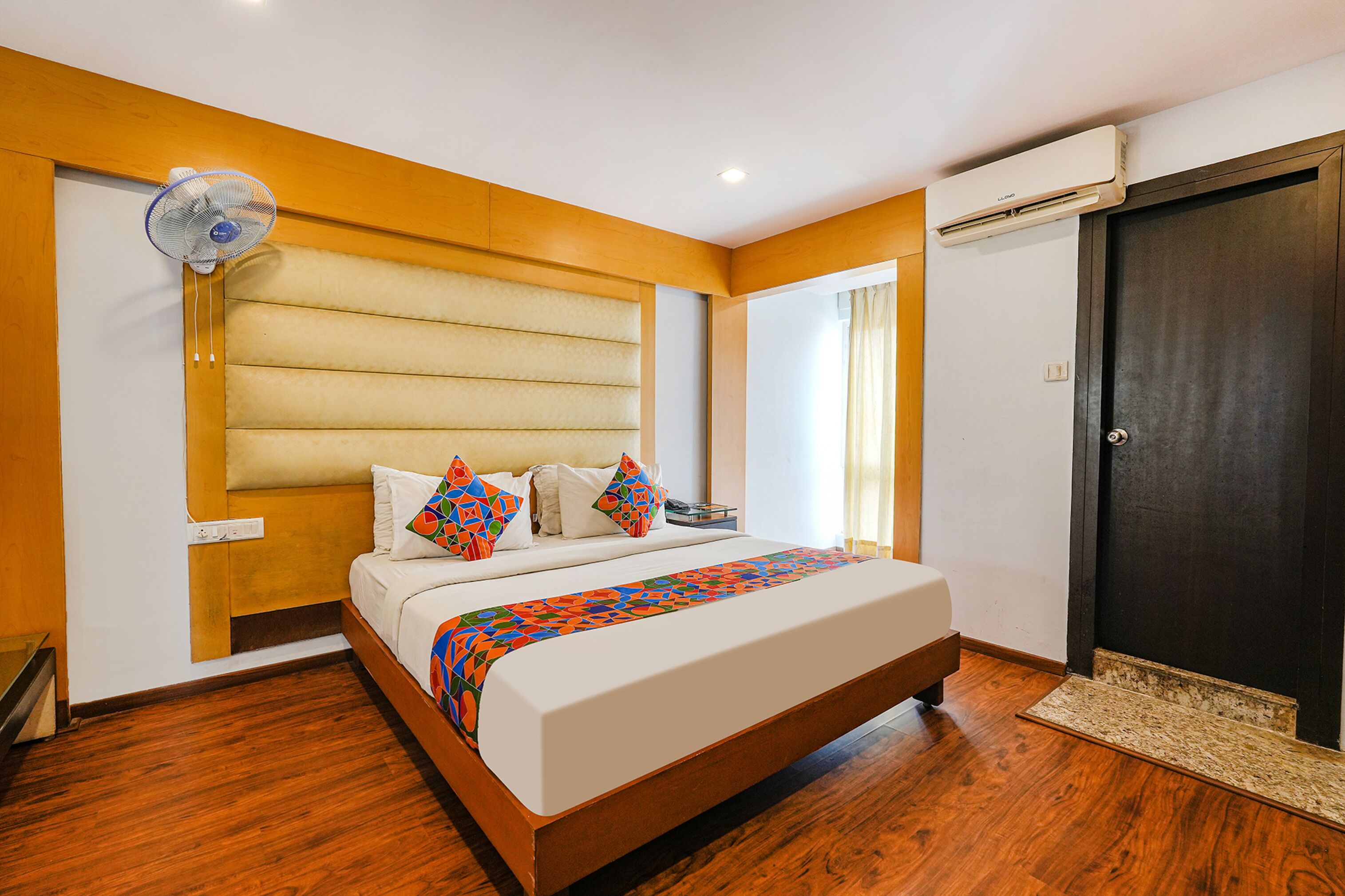 A bedroom with a bed, a standing fan, an air conditioning unit, and a dark door.