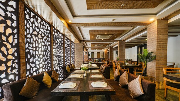 A restaurant with a long wooden table set for dining, surrounded by cushioned seating and a decorative wall. - FabHotel JVH (Bengaluru)