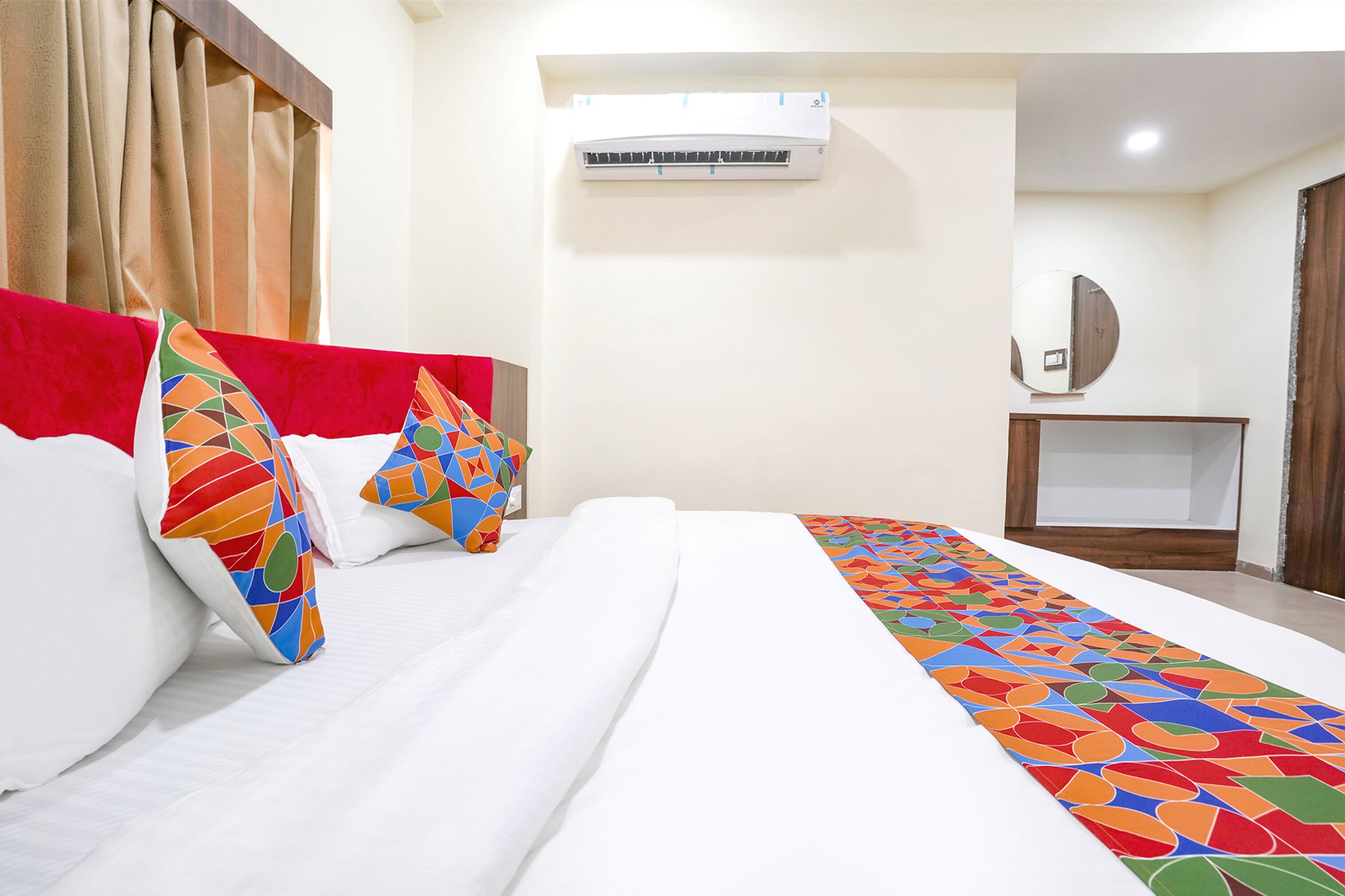 A hotel room with a bed, colorful pillows, a white bedspread, an air conditioning unit, a wooden headboard, a mirror, and a wooden cabinet.