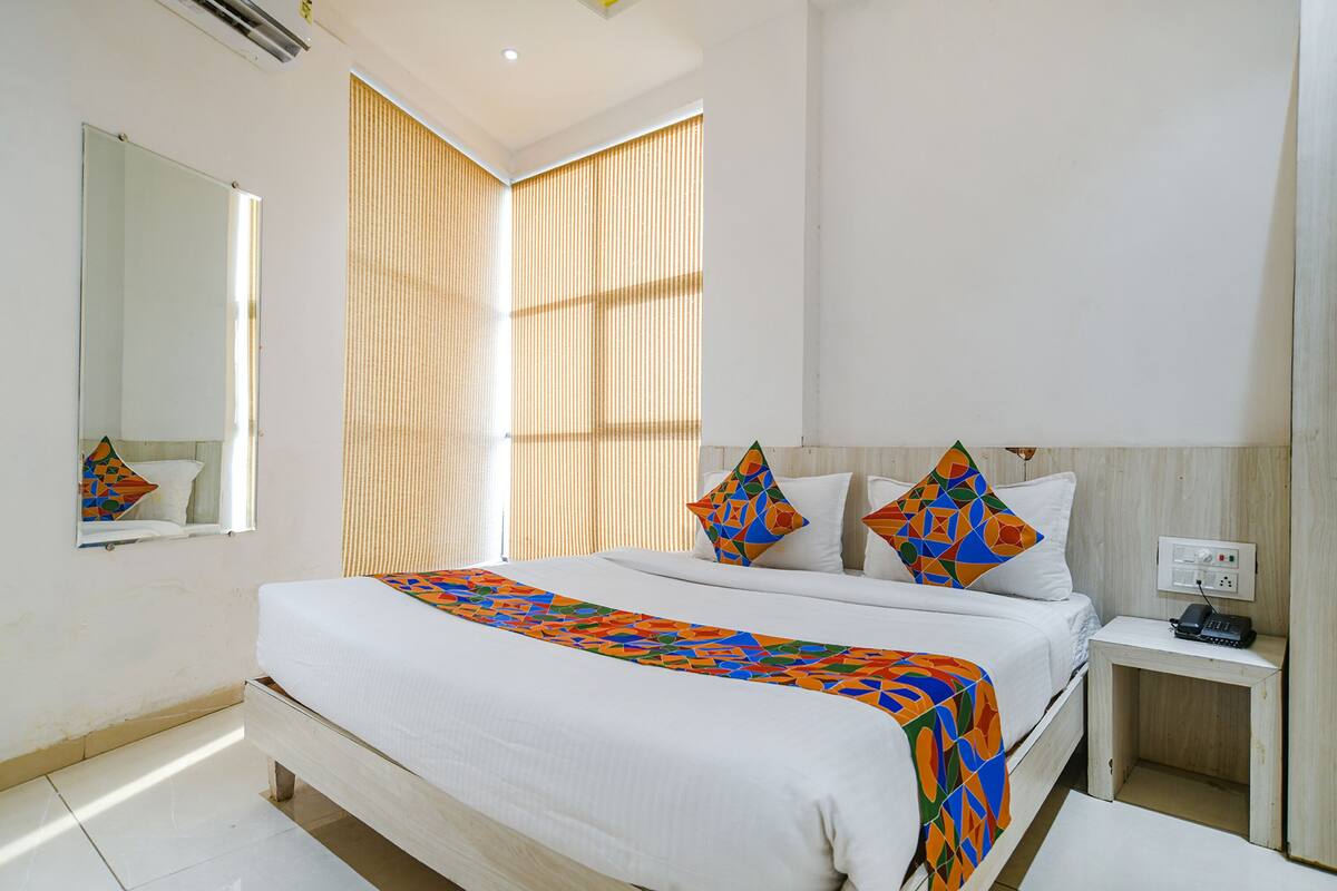 Habitación de hotel ordenada con una cama grande, una mesita de noche, un espejo y una ventana con persianas.