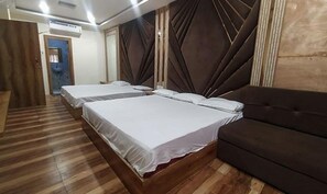 Habitación de hotel con dos camas, un sofá marrón oscuro, suelo de madera y un ventanal con cortinas.