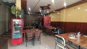A restaurant with wooden tables and chairs, a red beverage dispenser, and a patterned wall in the background. - Fabexpress Rudraksh Palace (Ujjain)
