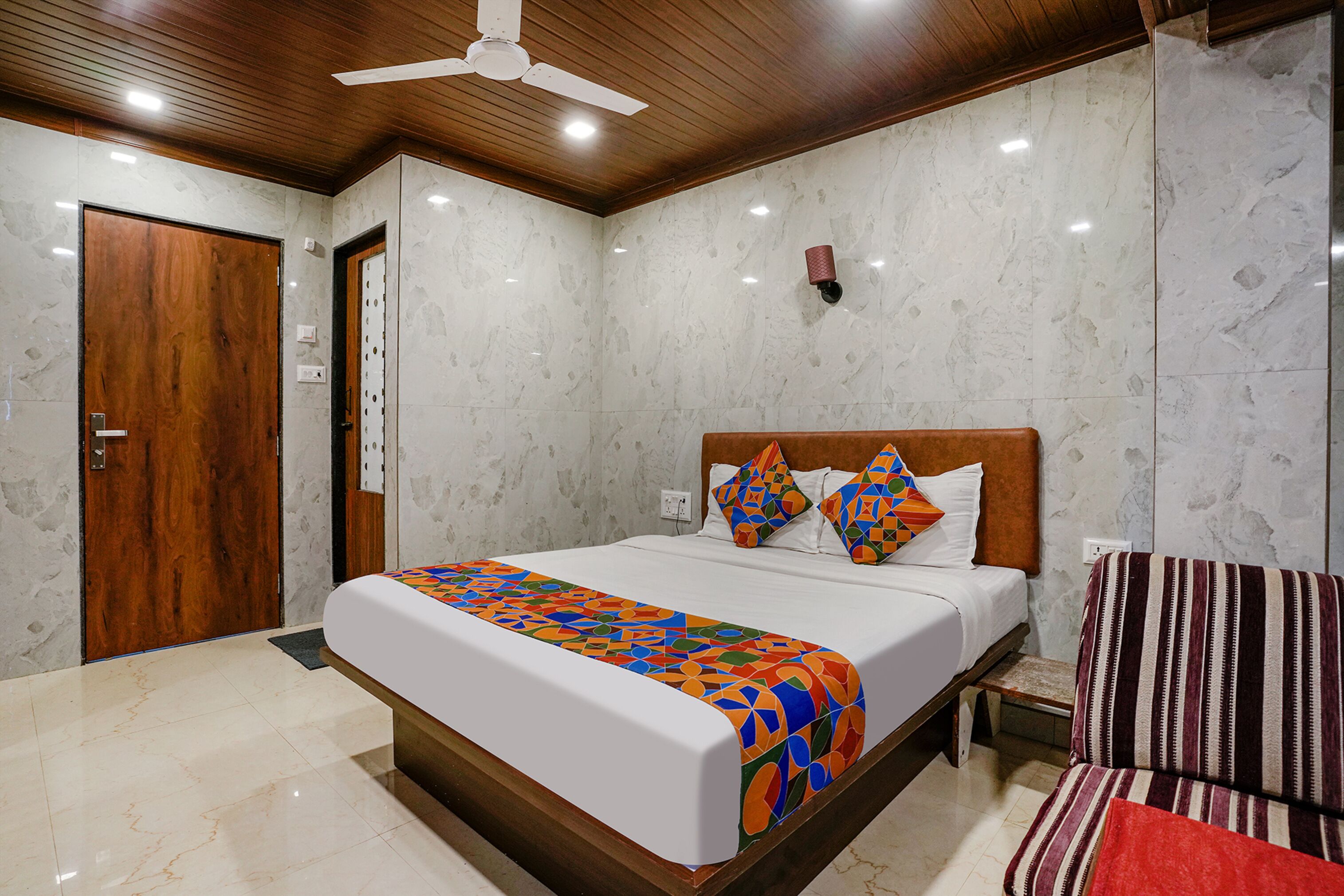 A hotel room with a wooden ceiling, a bed with a colorful bedspread, a striped armchair, and a marble wall.