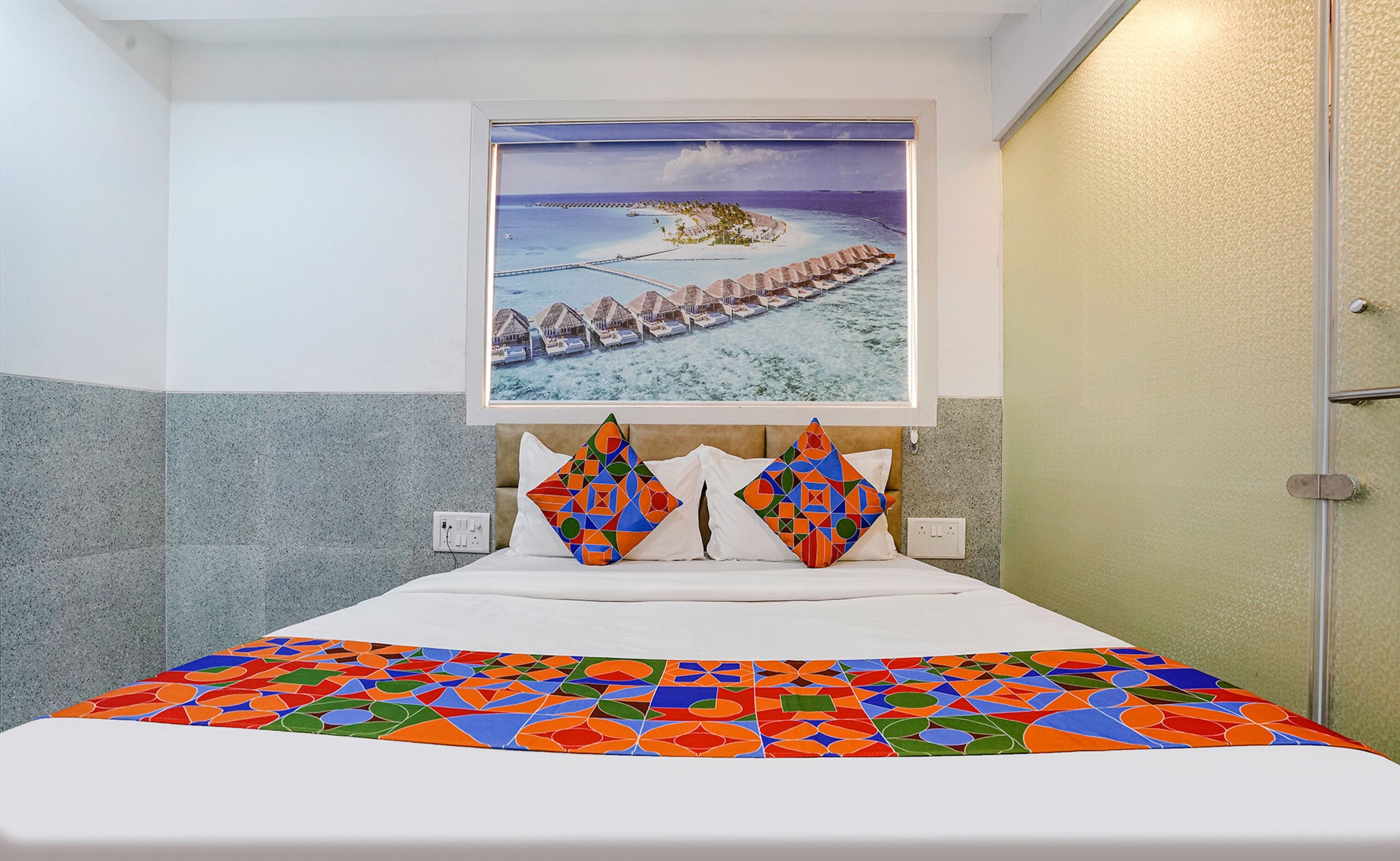 A hotel room with a bed, a colorful bedspread, a framed picture of a beach resort, and a door to the right.