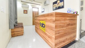 A reception desk with a wooden finish and a 'fab hotels' sign. There is a ceiling fan, a framed picture on the wall, and a small plant on the desk. - Fabhotel Living (Jaipur)
