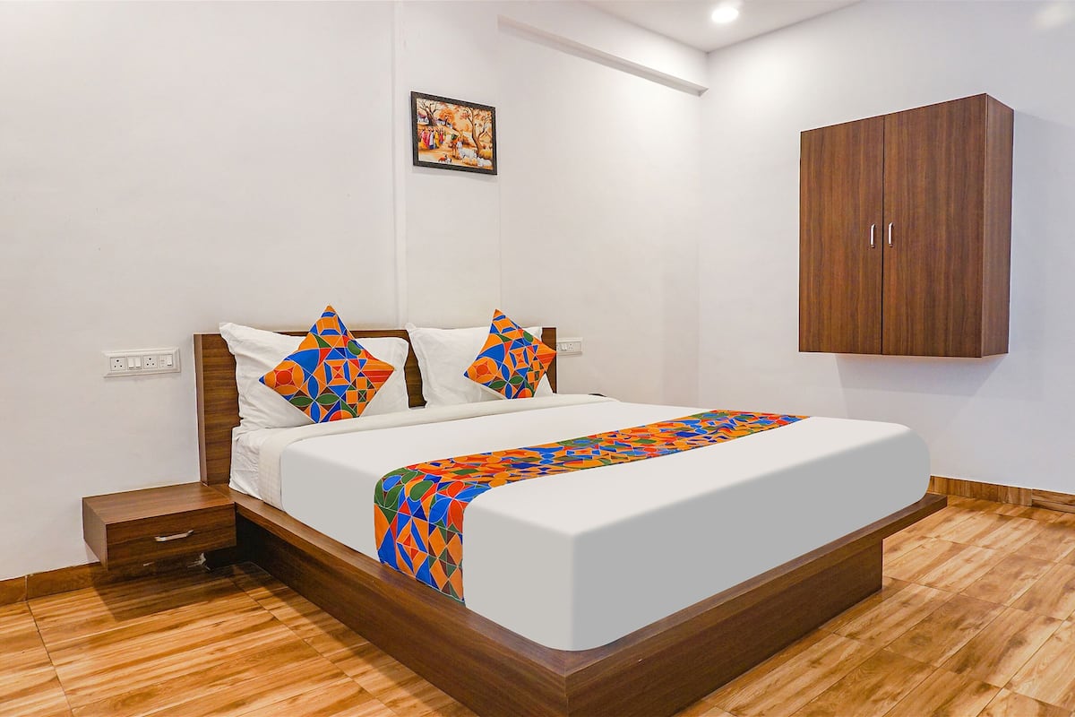 Una cama bien hecha con una manta de diseño geométrico y colores vivos, una mesita de noche de madera con cajón y un armario de madera frente a una pared blanca.