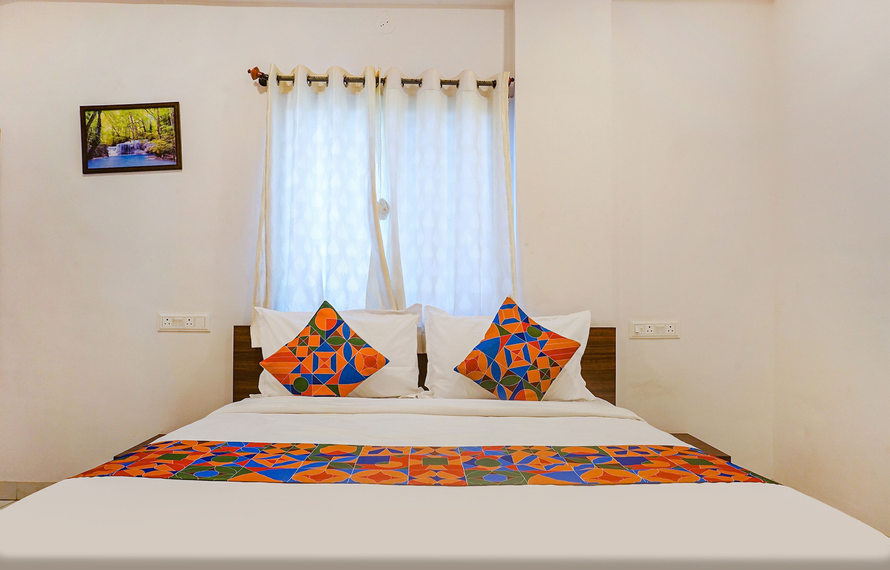 A neatly made bed with a colorful geometric-patterned bedspread, two pillows with a similar design, a framed picture of a natural landscape on the wall, and a window with white curtains.