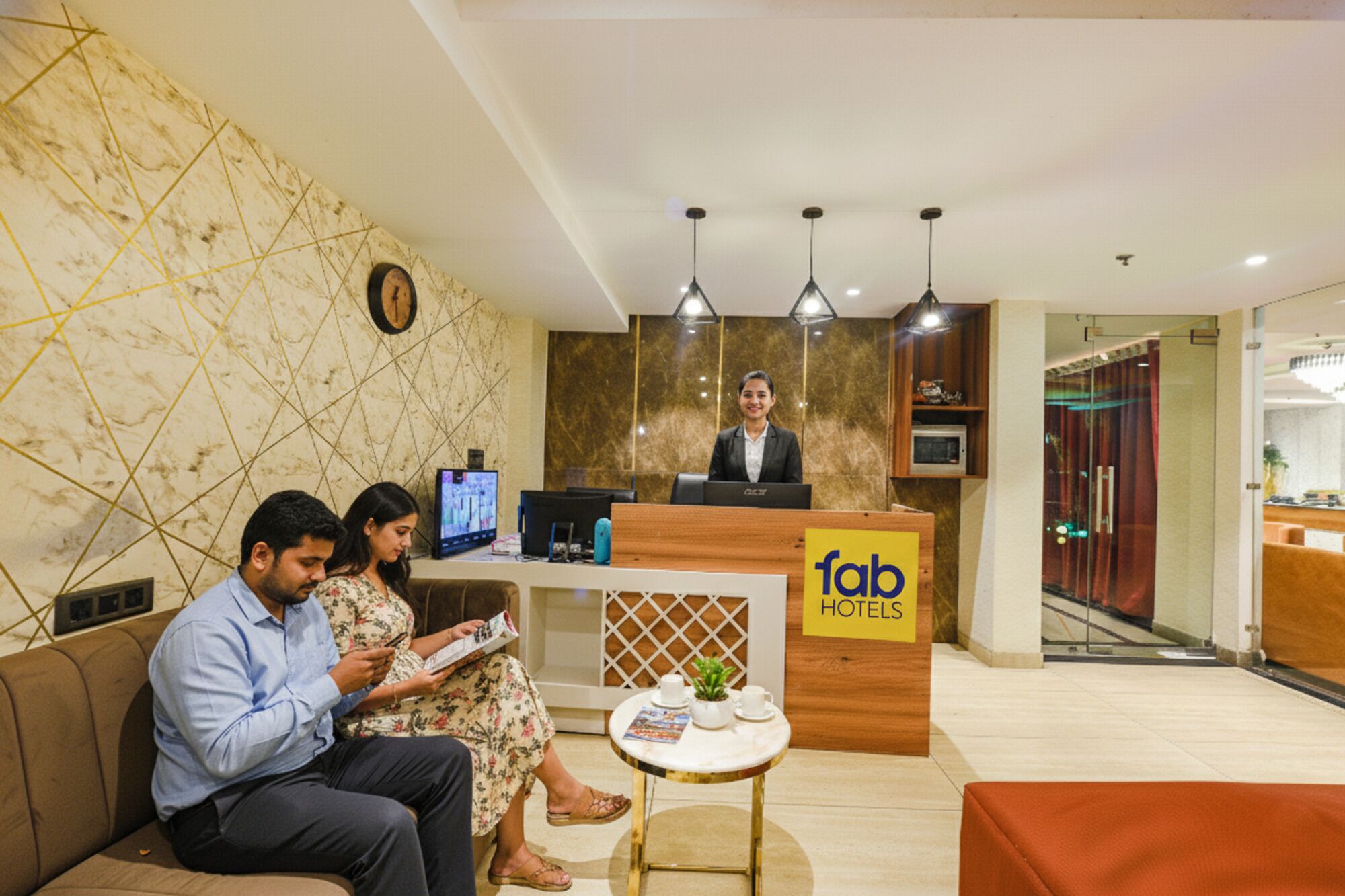 A hotel reception area with a receptionist, a reception desk, a clock, and a seating area with a small table.