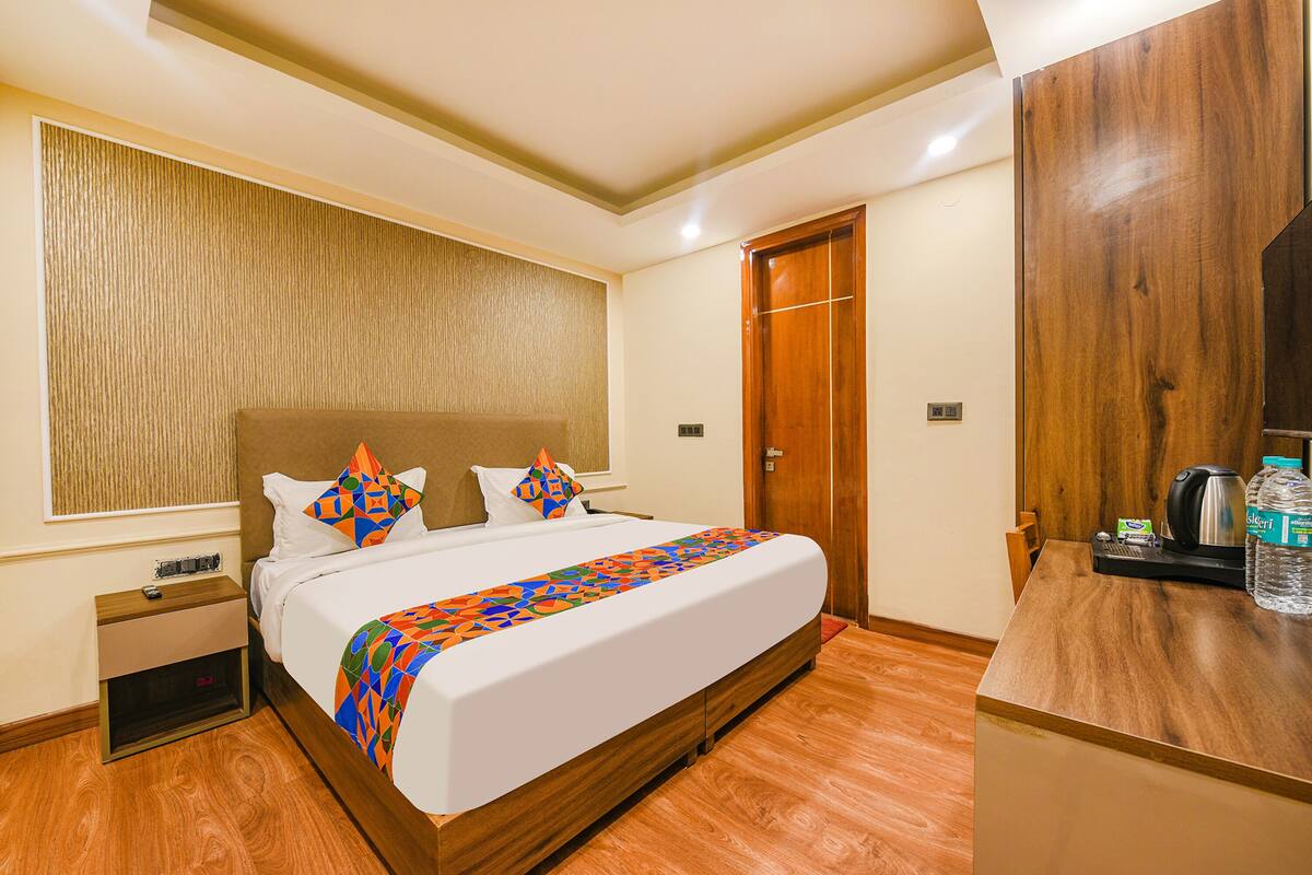 Habitación de hotel con una cama grande, cabecera de madera, mesita de noche, armario de madera y una pequeña mesa con hervidor y botella de agua.