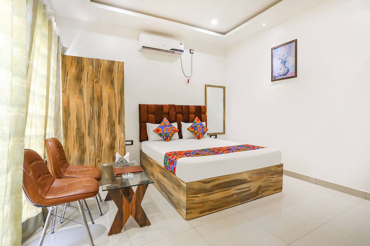 Une chambre d’hôtel avec un lit doté d’une tête de lit en bois, une table en verre, deux chaises marron, un petit bureau et un climatiseur fixé au mur.