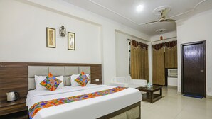 A hotel room with a large bed, a bedside table with a kettle, a small table with a bottle, and a door. - Fabhotel Corporate Stay (Noida)