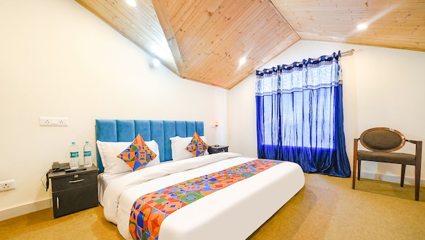 A bedroom with a wooden ceiling, a bed with a colorful bedspread, a nightstand, a chair, and a window with blue curtains. - Fabhotel Beer Castle (Theog)
