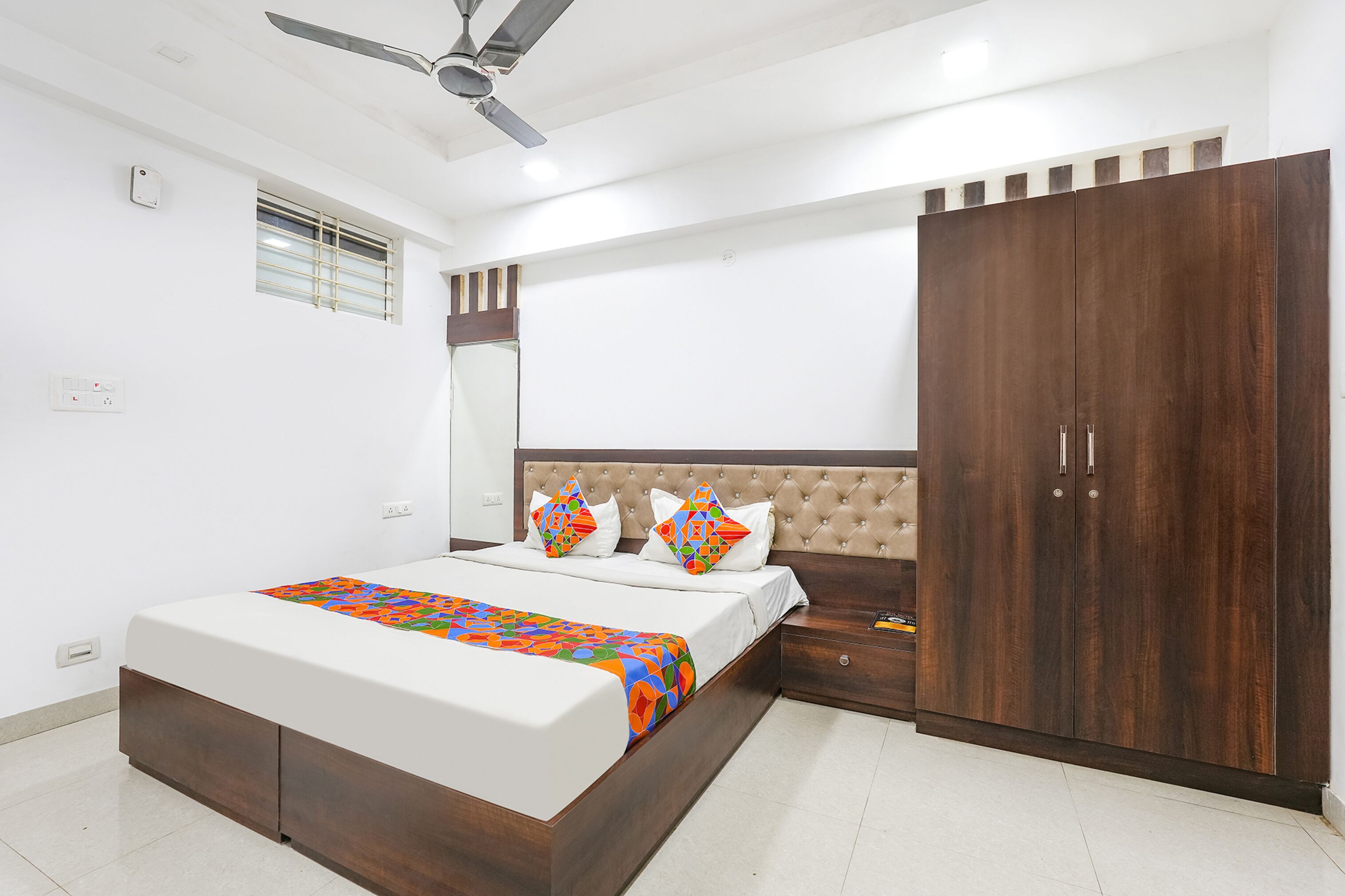 Une chambre à coucher comprenant un lit, une table de chevet, une armoire, un ventilateur de plafond et une fenêtre avec des stores.