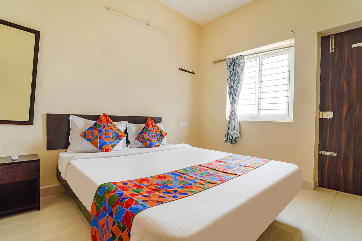 A neatly made bed with a colorful quilt, a bedside table, a window with curtains, and a wooden door.