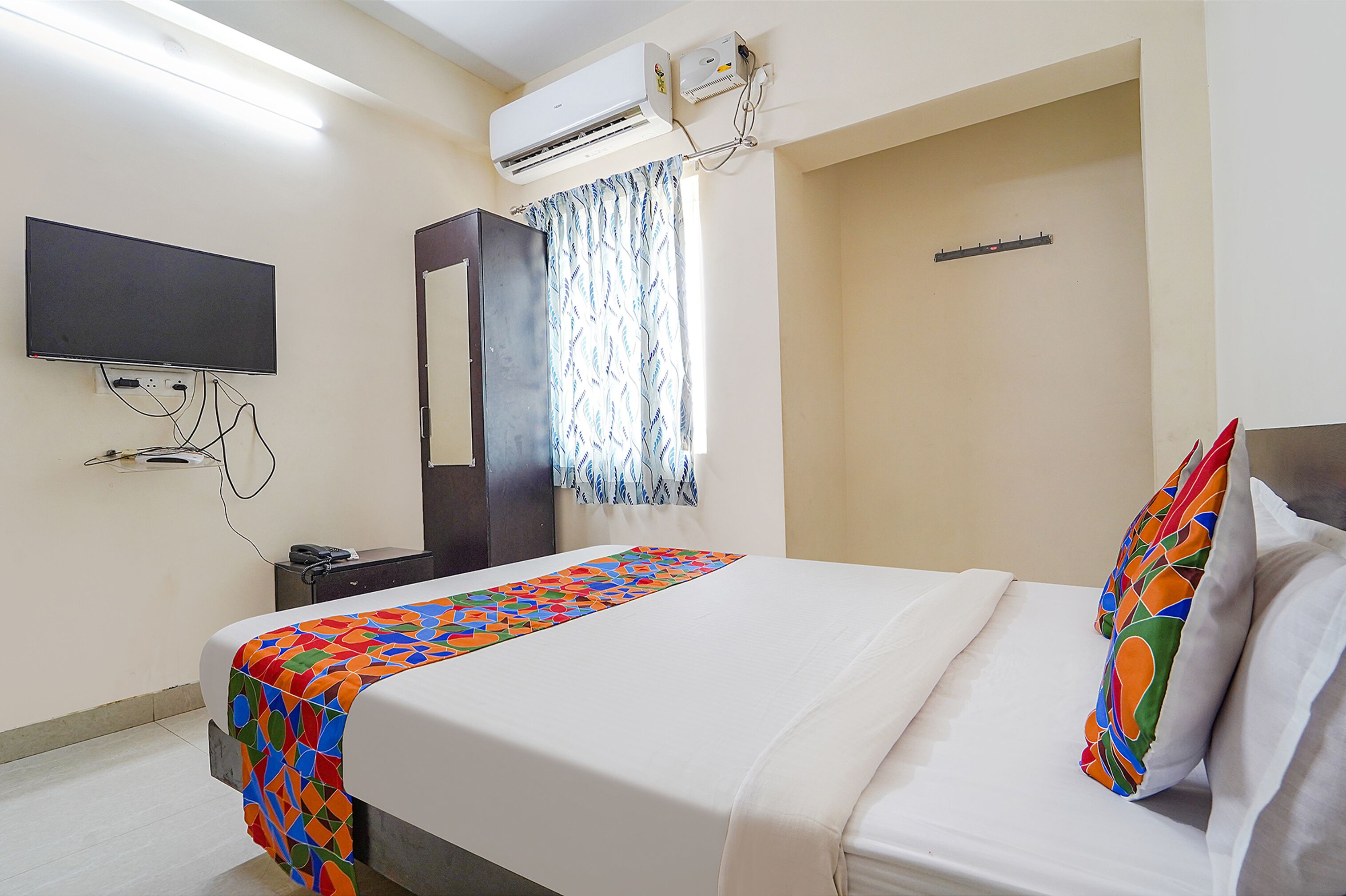 Une chambre d’hôtel équipée d’un lit, d’un téléviseur à écran plat fixé au mur, d’un appareil de climatisation et d’une fenêtre avec des rideaux à motifs.