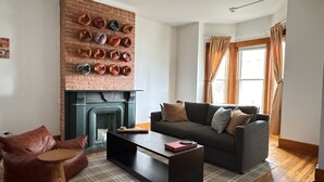 Un salón con chimenea de ladrillo, un sofá gris oscuro, poufs de cuero y un tapete escocés.