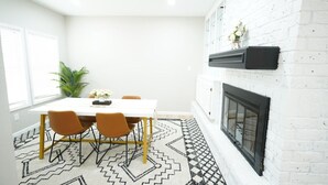 Un comedor con una mesa blanca, cuatro sillas, un tapete estampado, una pared de ladrillo blanco y una chimenea negra.