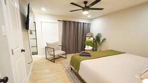 Un dormitorio con una cama, una silla, un ventilador de techo, una ventana con cortinas y una planta.