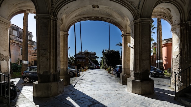 An arched corridor with stone columns, leading to a street with parked cars and buildings in the background. - Rua de Mar Portici (Portici)