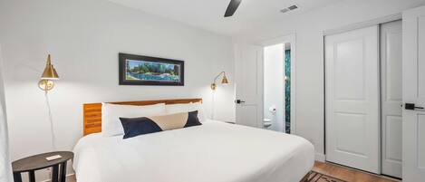 A neatly arranged hotel room with a bed, a ceiling fan, a framed picture on the wall, and a door leading to another room.