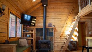 Un accogliente interno di una baita con camino in pietra, pareti in legno, TV a muro e una scala in legno.