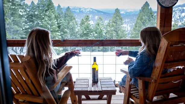 Due persone sedute su sedie di legno attorno a un tavolo di legno, con una bottiglia di vino, con vista su un paesaggio montuoso attraverso una finestra a griglia.