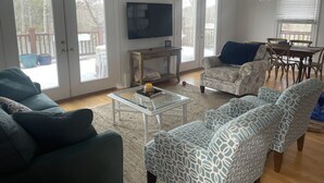 Ein Wohnzimmer mit einer Couch, einem Sessel und einem Couchtisch. An der Wand ist ein Fernseher angebracht und es gibt einen Esstisch mit Stühlen.