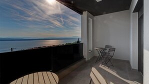 A balcony with a view of a lake and mountains, a small table with chairs, and a sun shining through the clouds.