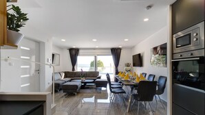 A modern living room with a sofa, dining table, and chairs. There is a kitchen area with a microwave and oven.