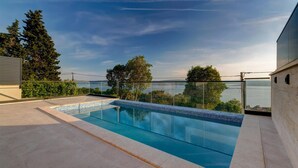 A modern outdoor swimming pool with clear blue water, surrounded by a glass balustrade, set against a backdrop of trees and a distant view of the sea.