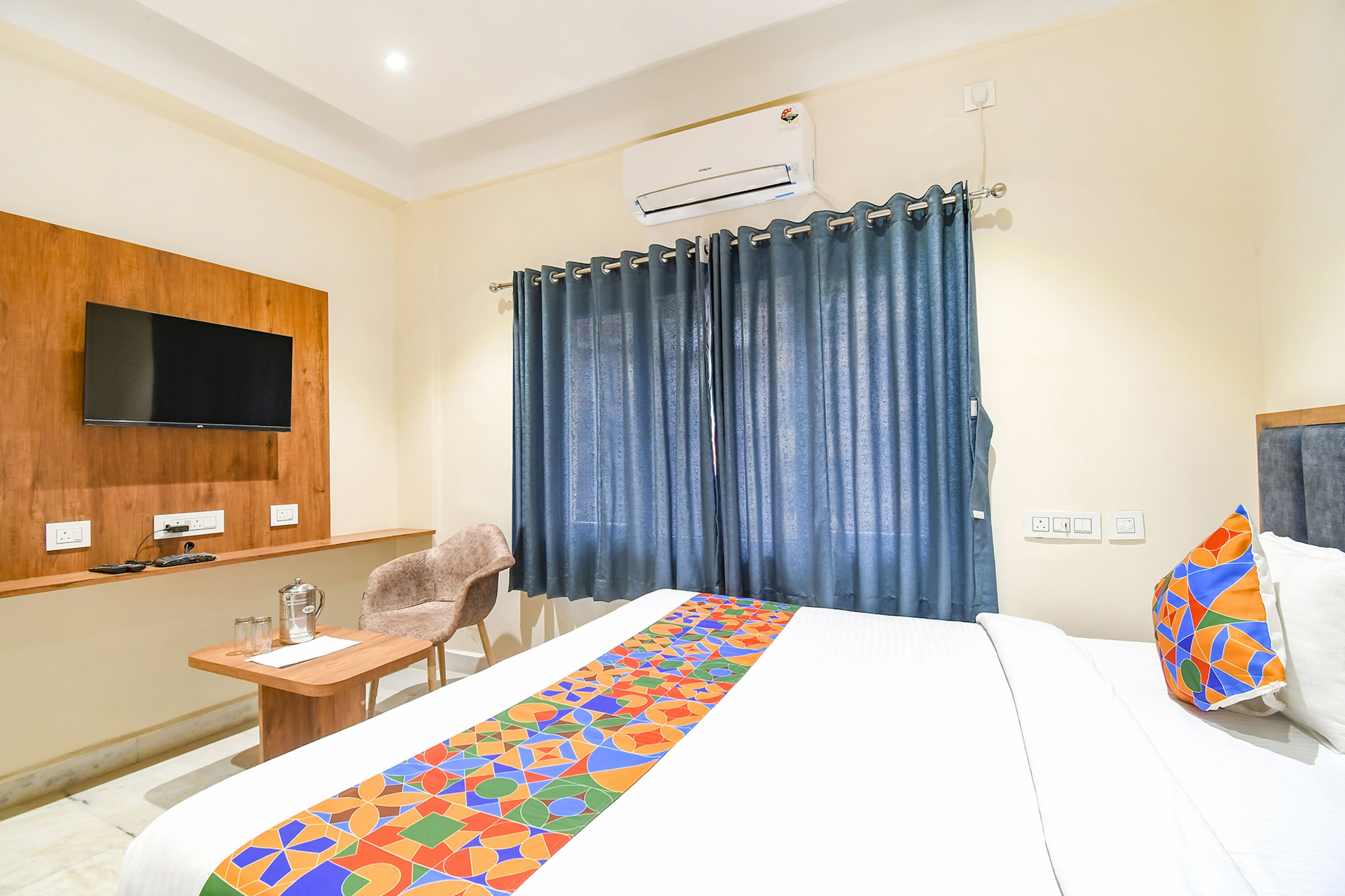 A hotel room with a bed, a TV mounted on the wall, a desk with a chair, and a window with curtains.